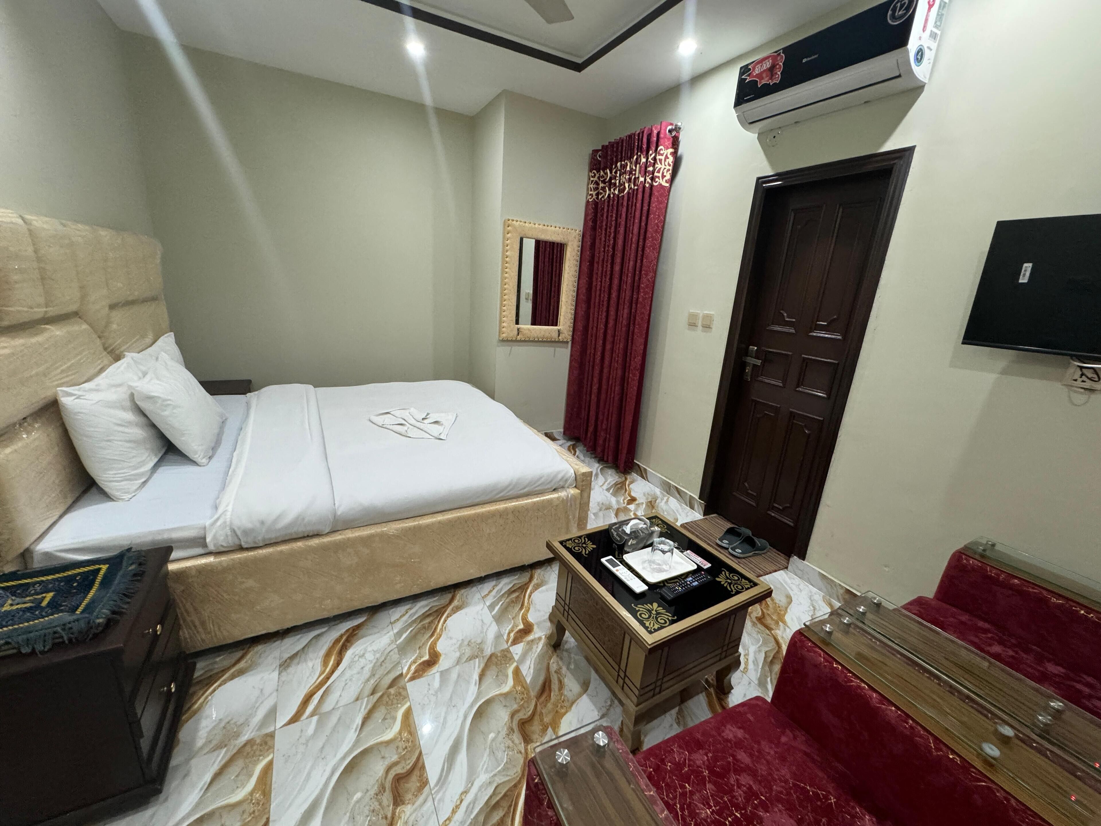 A hotel room with a bed, a small bedside table with a phone and charger, a red chair, a wooden cabinet, and a TV mounted on the wall.