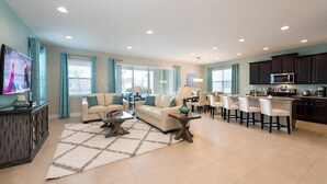 A spacious living room with a large TV, two sofas, a coffee table, and a dining area with chairs and a kitchen in the background.