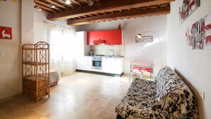 Un salon chaleureux avec un canapé à motifs, une étagère en osier et un coin repas comprenant une table et des chaises. On aperçoit au fond une cuisine avec des placards rouges et un fourneau.