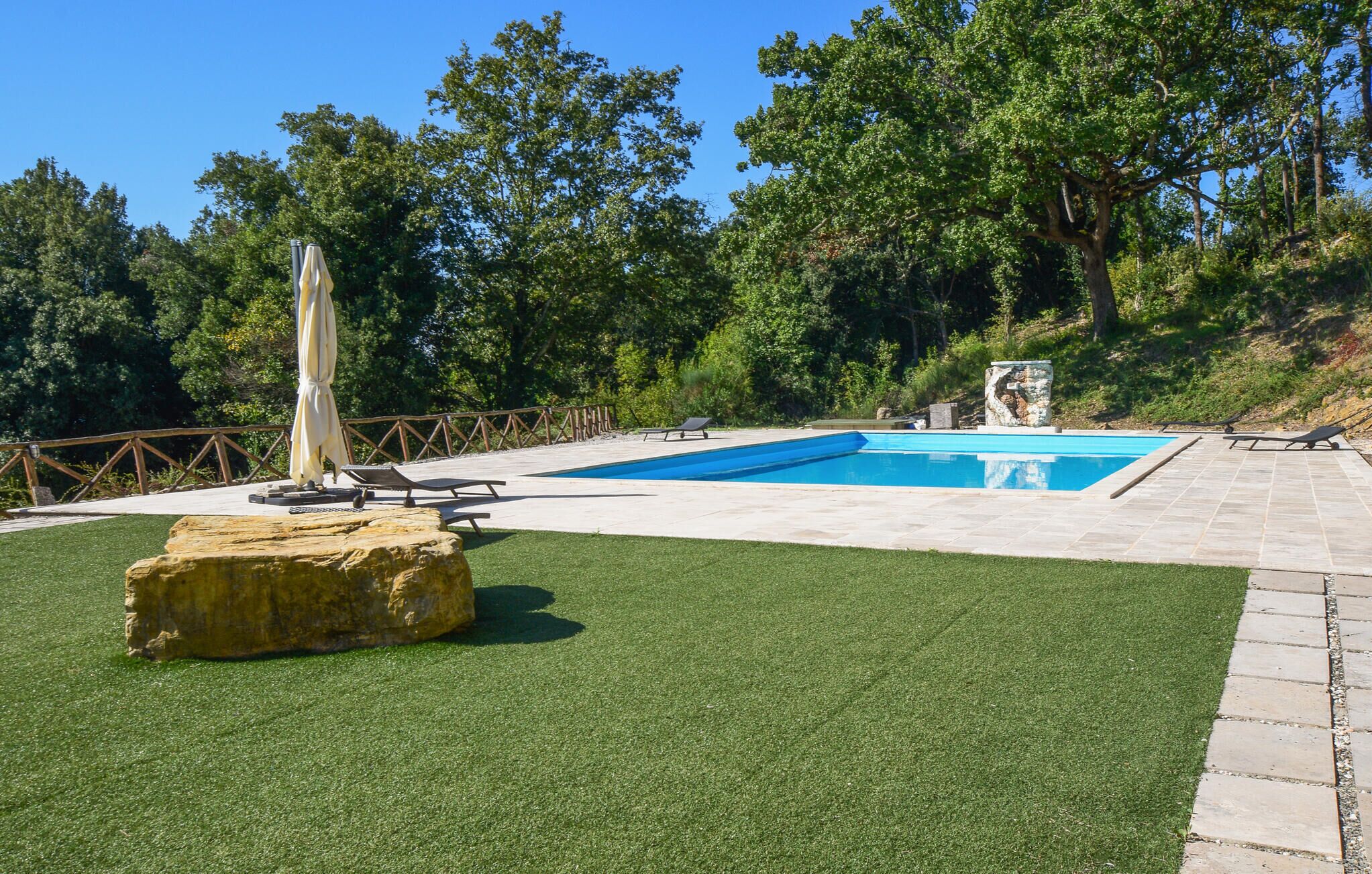 Une piscine dotée d’une plage en pierre, entourée de gazon synthétique et d’arbres.