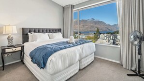 Une chambre avec un grand lit, une table de chevet, une lampe, un ventilateur et une vue sur les montagnes et un lac, visible à travers une grande fenêtre.