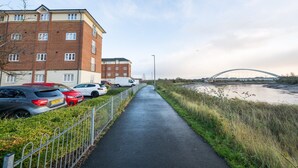 Ein Weglauf entlang eines Flusses, im Hintergrund eine Brücke, eine Reihe geparkter Autos und ein mehrstöckiges Gebäude mit Backsteinfassade.