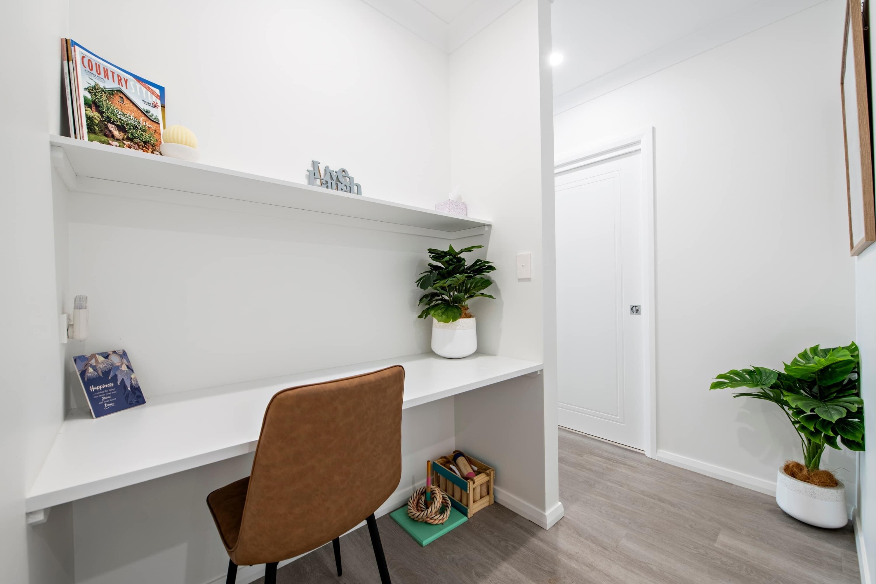 Una habitación limpia y blanca con un escritorio, una silla y estantes con libros y plantas.