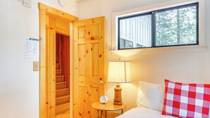 A bedroom with a wooden door, a bedside table with a lamp, a checkered pillow, and a window with a view of trees. - < 2 Blocks to Lake Tahoe: Family Home Near Skiing (Homewood)