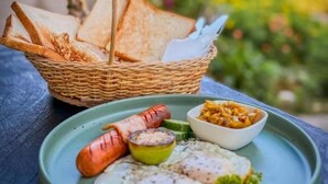 A plate with eggs, sausages, and a side of greens, accompanied by a basket of bread and a small bowl of sauce, set on a table outdoors. - Tera Ella Boutique Hotel by SANS (Ella)