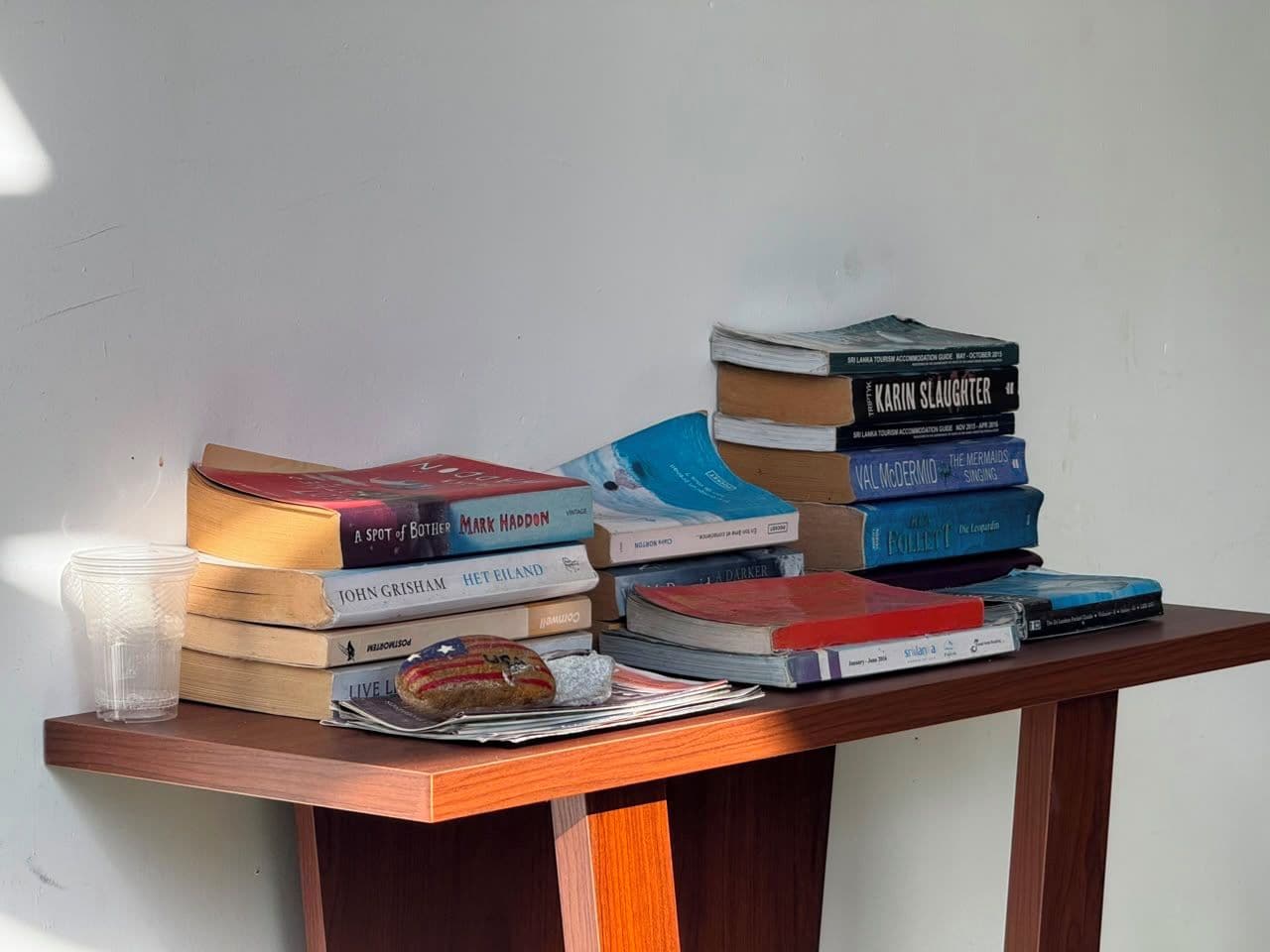 A stack of books on a wooden table with a clear plastic cup and a pastry.