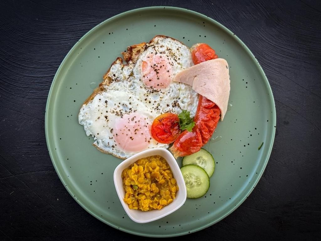 Un plato con un huevo frito, rodajas de tomate, pepino y un recipiente pequeño con un condimento amarillo.