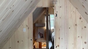 Een smalle houten gang met een schuine balken plafond en een deur naar een kamer met een bed en een lijst met een afbeelding erop.