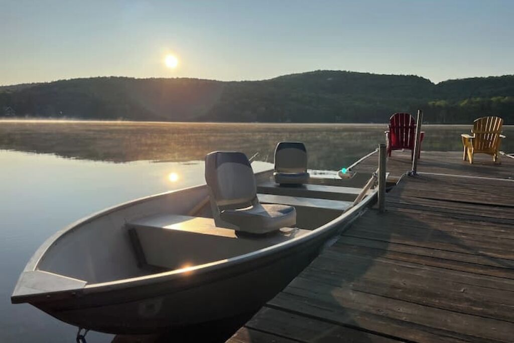 Een boot afgemeerd aan een houten steiger, met stoelen en uitzicht op een rustig meer en heuvels op de achtergrond.