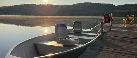 Een boot afgemeerd aan een houten steiger, met stoelen en uitzicht op een rustig meer en heuvels op de achtergrond.