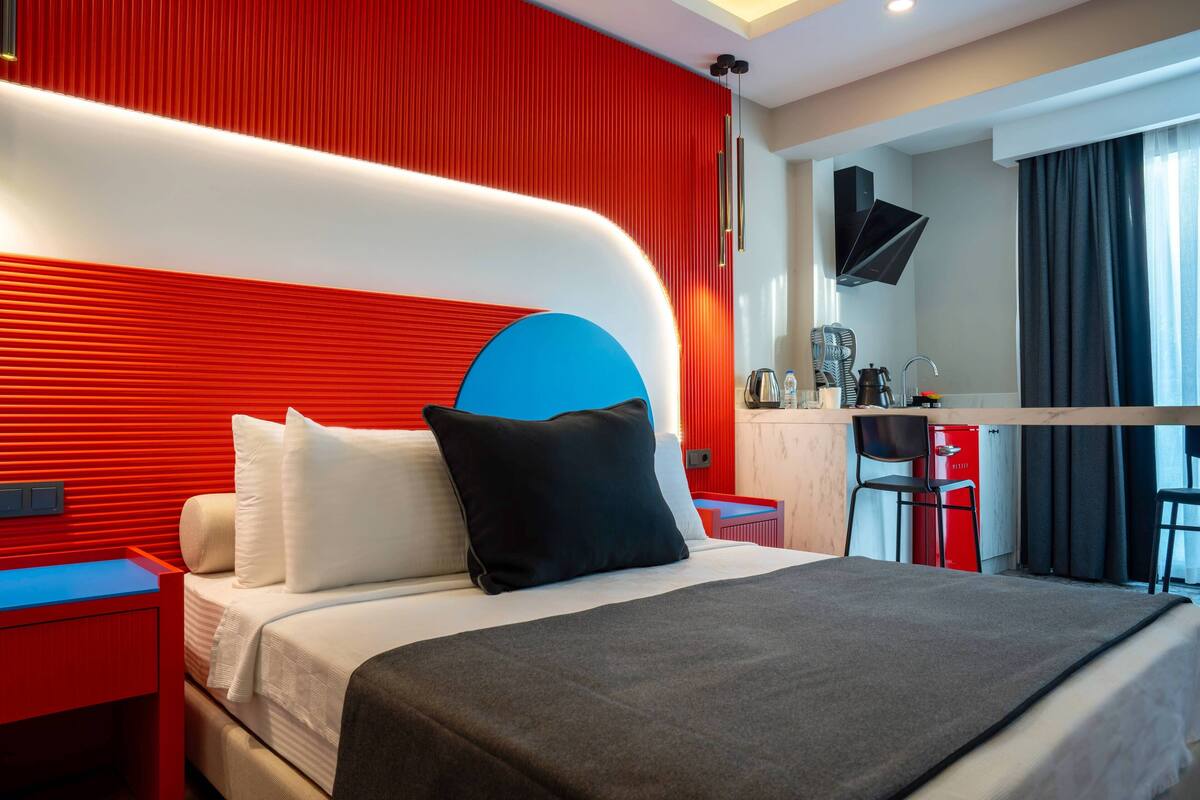 A modern hotel room with a red and white color scheme, a bed with white and black bedding, a desk with a chair, and a flat-screen TV mounted on the wall.