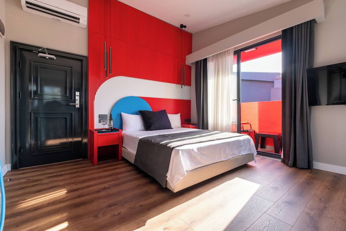 A modern hotel room with a large bed, a red accent wall, a black door, and a sliding glass door leading to an outdoor area.