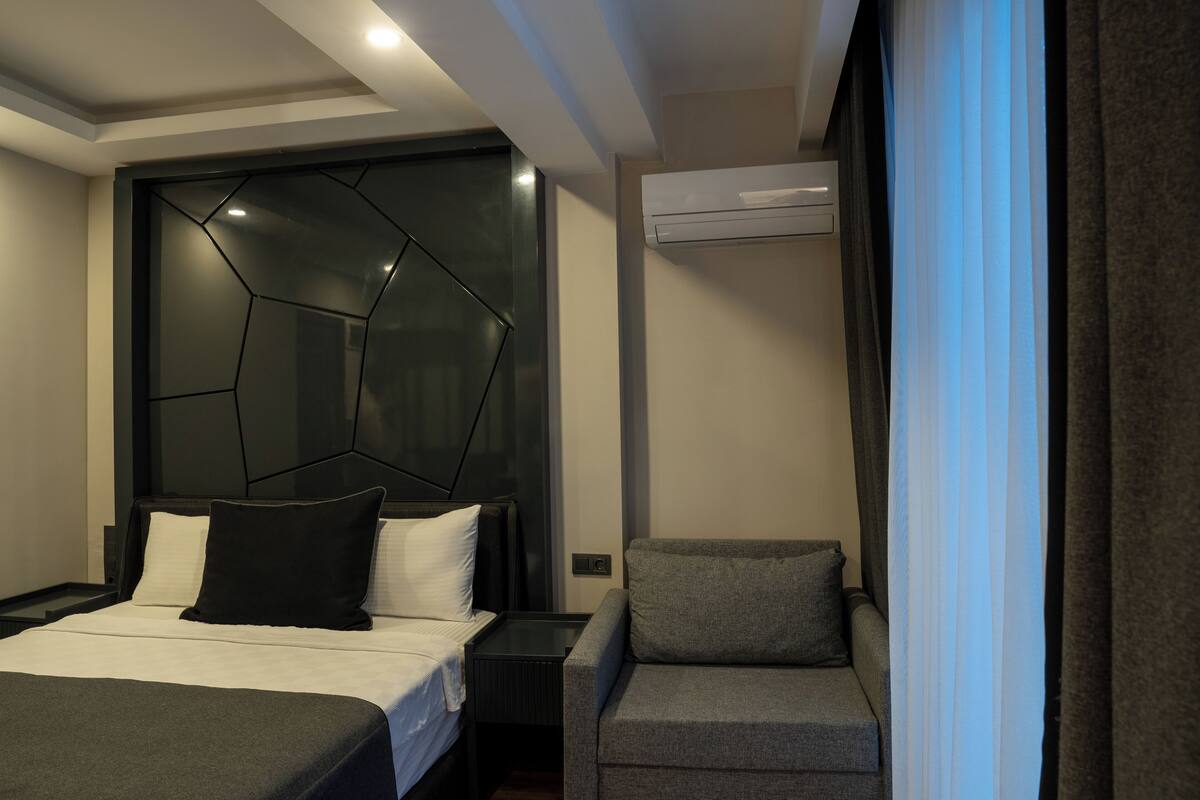 A modern hotel room with a large bed, a grey sofa, a geometric-patterned headboard, and an air conditioning unit.