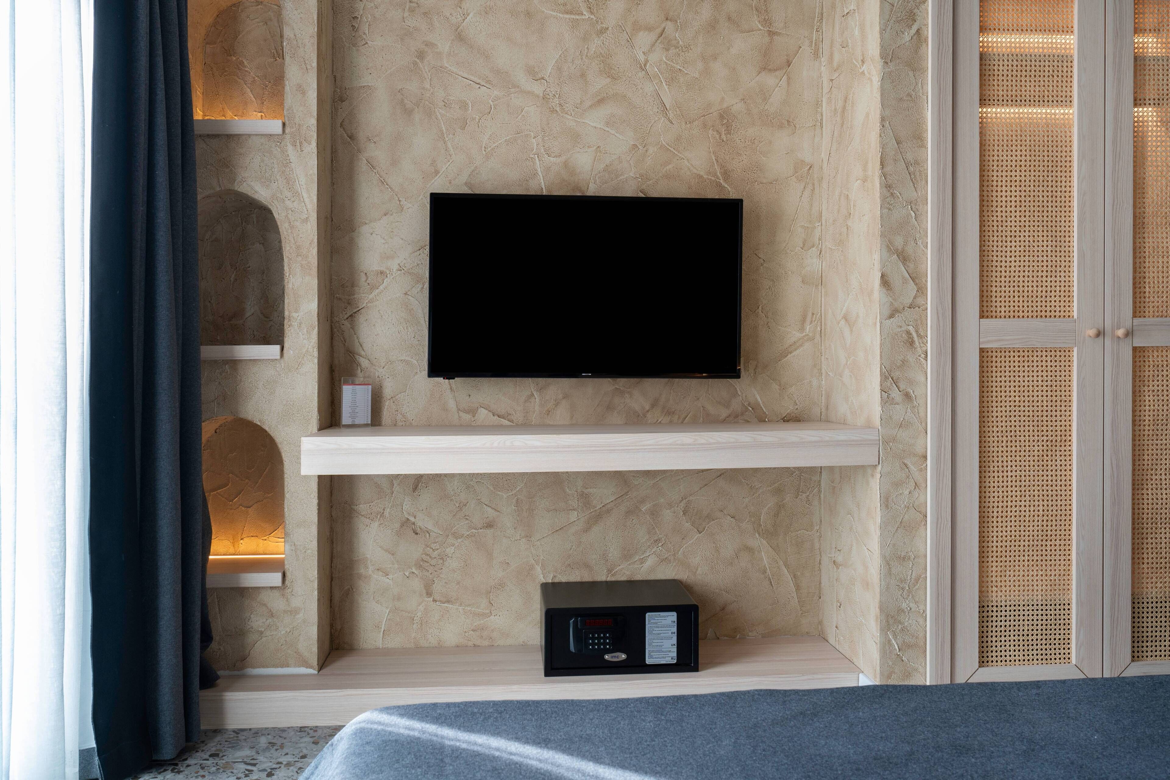 Une chambre d’hôtel moderne équipée d’un téléviseur à écran plat fixé sur une étagère en bois, d’un petit panneau de commande en dessous et d’une grande fenêtre avec des rideaux.