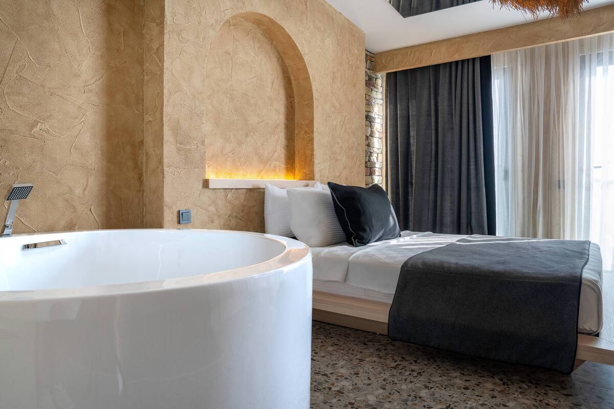A modern hotel room with a large bathtub, a bed with a dark blanket, and a stone wall.