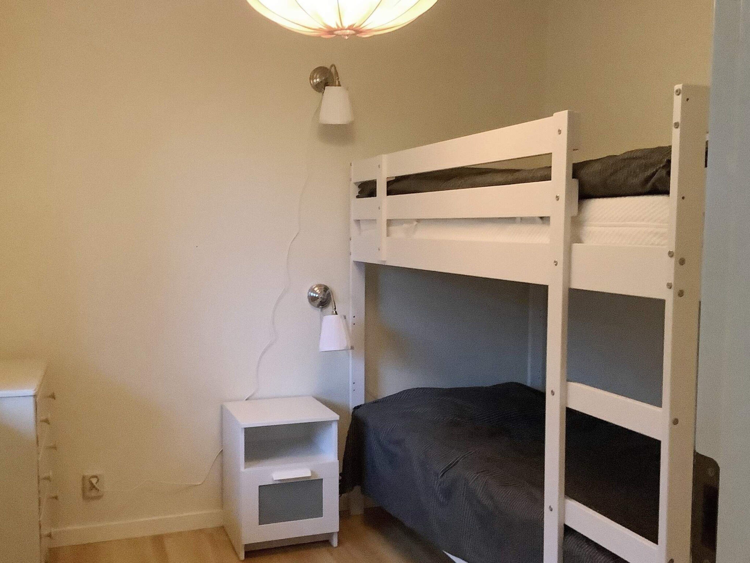 A bunk bed with a nightstand and a lamp, in a room with plain walls and a light fixture.