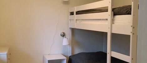 A bunk bed with a nightstand and a lamp, in a room with plain walls and a light fixture.