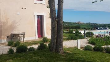 A building with a red door, a table and chairs outside, and a view of a pool and greenery in the background.