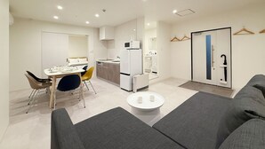 Ein modernes, helles Wohnzimmer mit Sofa, Esstisch und Stühlen, einer Küchenzeile mit Kühlschrank sowie einem Badezimmer mit Spiegel und Handtuchhalter.