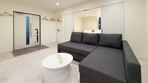 Ein modernes Hotelzimmer mit einem grauen Modulsofa, einem weißen Couchtisch und einem großen Spiegel. Eine Tür mit blauer Streifen sowie ein Kleiderständer mit Kleiderbügeln sind ebenfalls vorhanden.