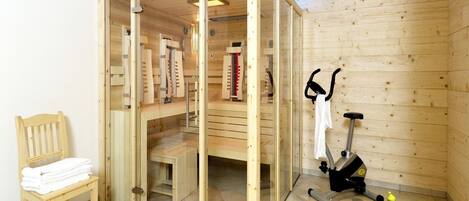Un cuarto de sauna con paredes de madera y puerta de vidrio, una silla de madera con toallas, una colchoneta azul para ejercicios y una bicicleta estática.