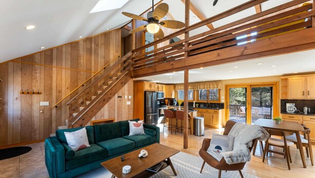 A spacious living area with a green sofa, a wooden coffee table, and a dining table with chairs. A staircase leads to an upper level, and the kitchen is visible in the background.