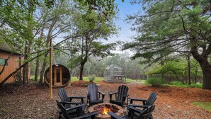 A fire pit surrounded by four Adirondack chairs in a forested area with a wooden cabin and a glass greenhouse in the background.