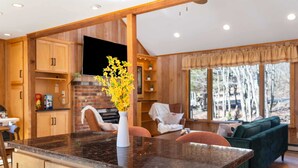 A kitchen with a granite countertop, wooden cabinets, a TV mounted on the wall, and a window with a view of trees.