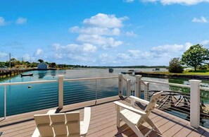 Una terraza con sillas que da a una masa de agua tranquila, un muelle y una orilla lejana.