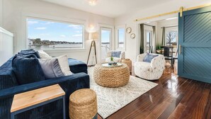 A spacious living room with a large window, a blue sofa, a round woven ottoman, and a white armchair. - Oceanfront Dock Escape ~ Kayaks & Paddleboards (Gloucester)