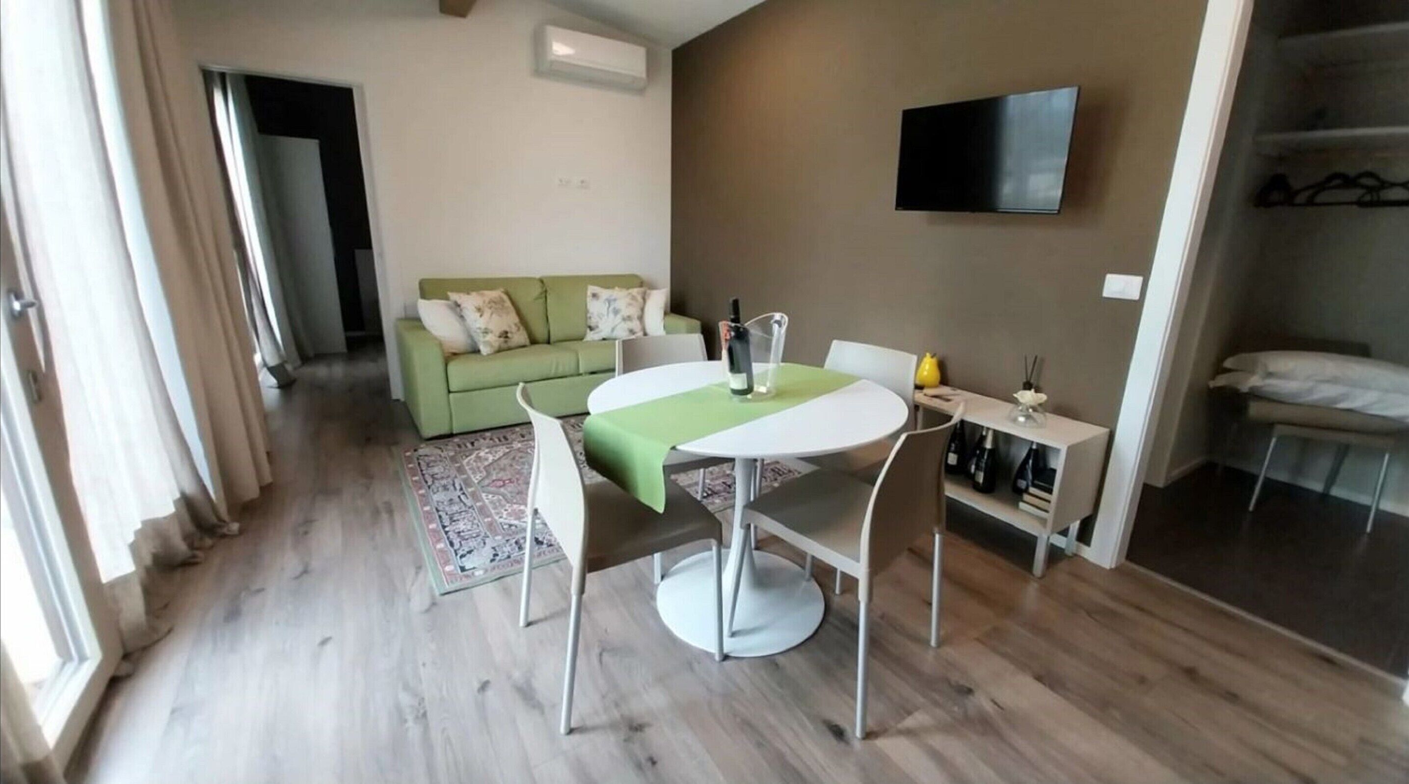 A modern hotel room with a dining area, a green sofa, a TV mounted on the wall, and a small table with a bottle of wine.