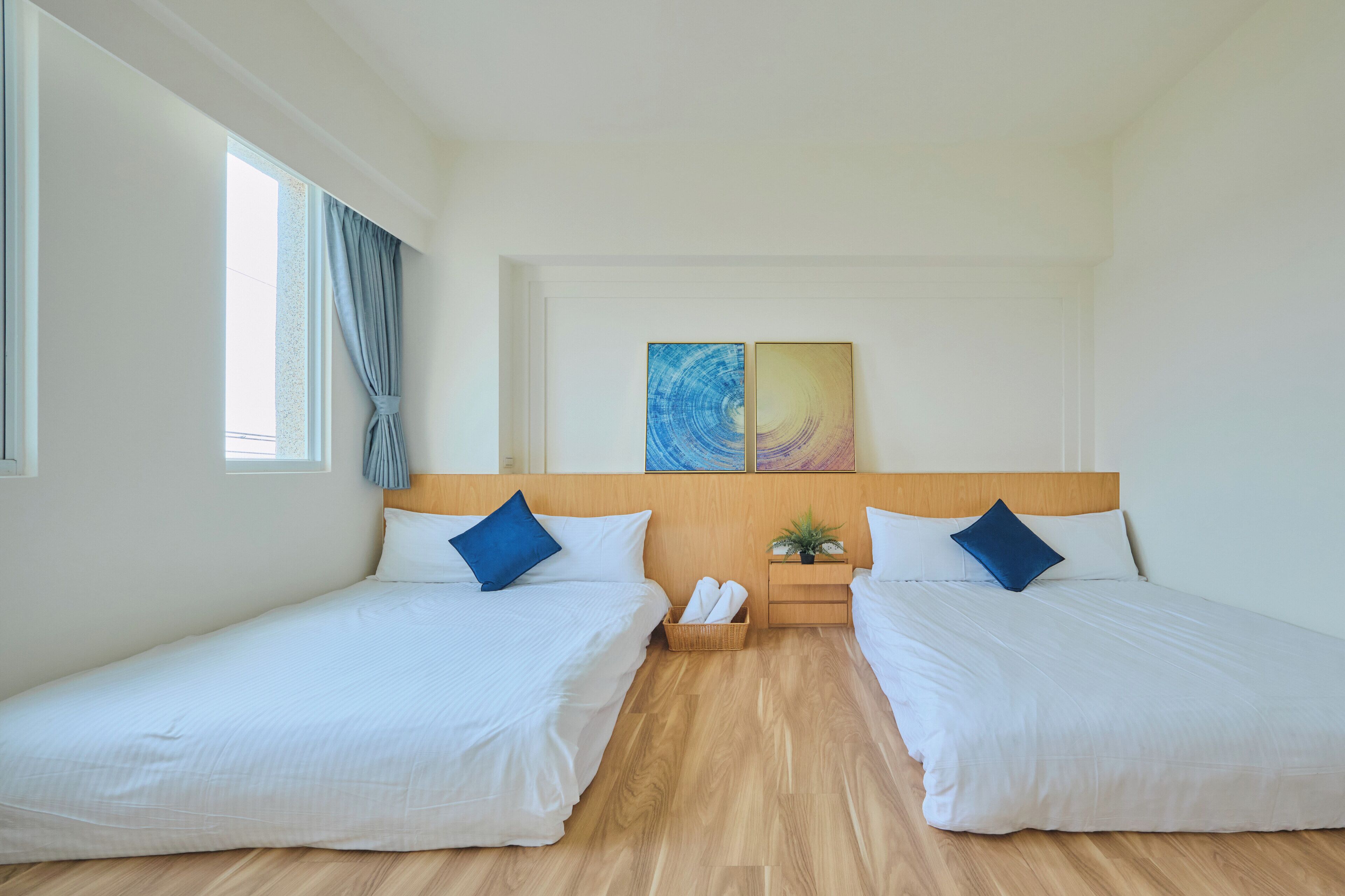 A hotel room with two single beds, a wooden headboard, a small plant, and a window with curtains.