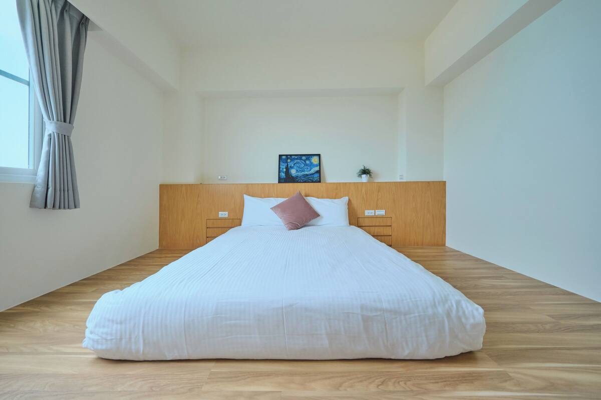 A neatly made bed with a wooden headboard, a single pink pillow, and a painting above it.