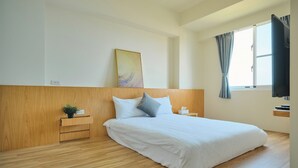 Una habitación de hotel moderna con cabecera de madera, una cama grande con ropa de cama blanca, una mesita de noche pequeña con una planta y una ventana con cortinas.