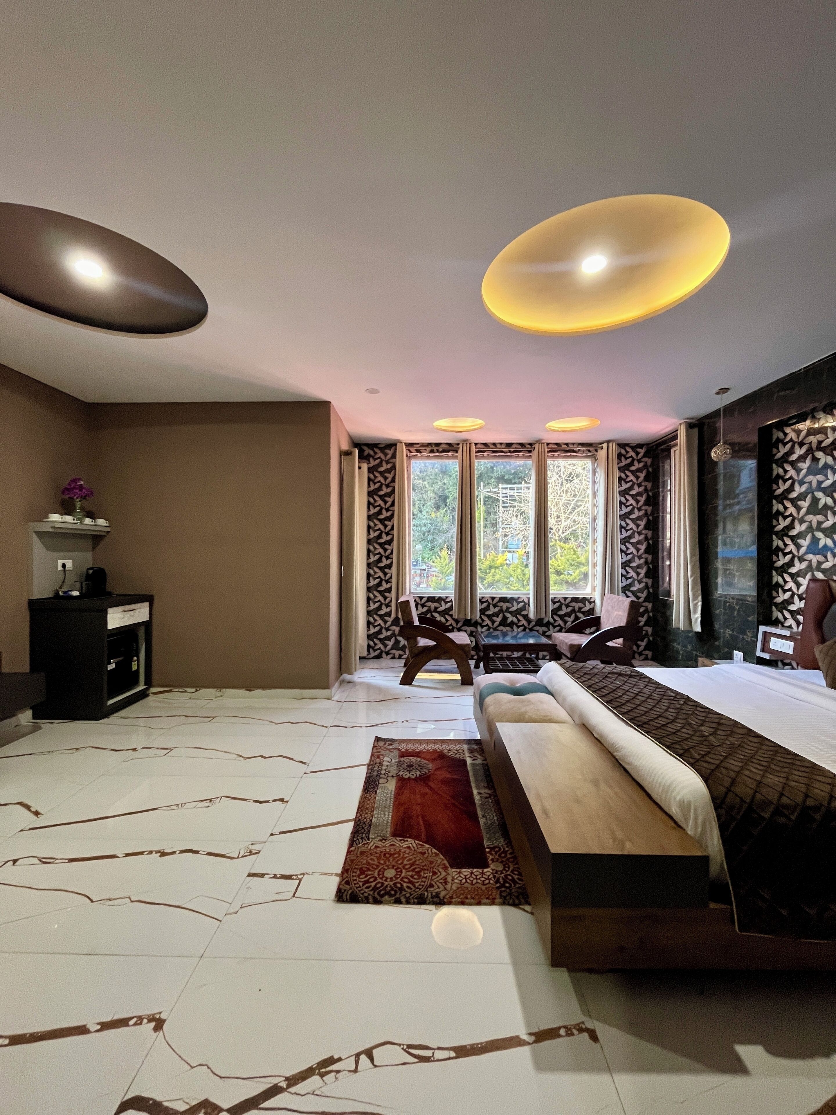 Ein modernes Hotelzimmer mit einem großen Bett, einem Schreibtisch und einem Stuhl. Es gibt Fenster mit gemusterten Vorhängen, eine Deckenleuchte und einen dekorativen Teppich.