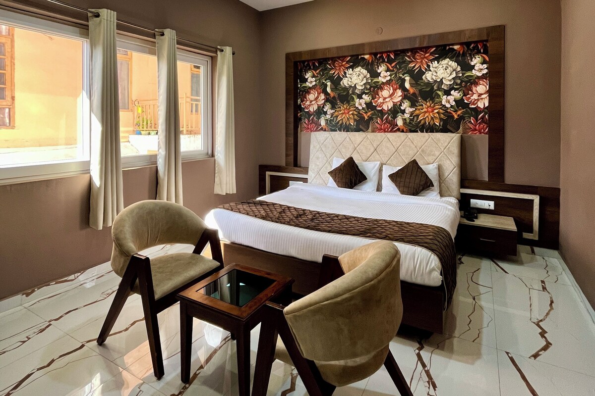 A hotel room with a bed, two armchairs, a small table, a nightstand, and a large floral artwork on the wall.