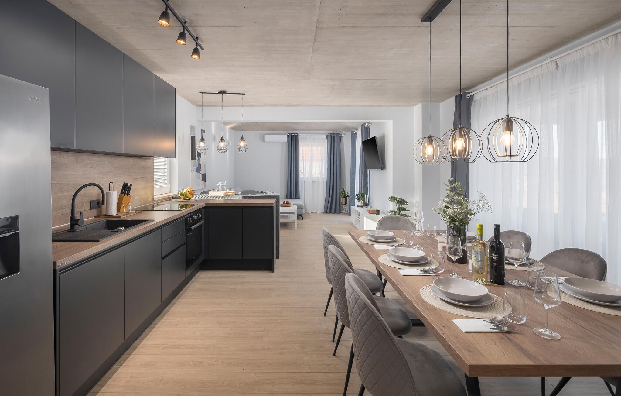 Una cocina moderna con una isla central, comedor con una mesa de madera preparada para seis personas, y un espacio de sala de estar con televisor y asientos.