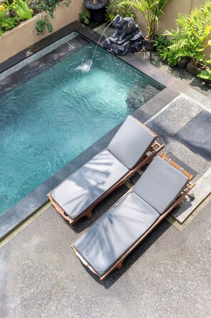 A rectangular pool with clear water, surrounded by a stone border. There are two wooden lounge chairs with grey cushions.