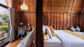 A wooden room with a bed, a desk, and a chair. There is a window with curtains and a view of the outside. - Lumbung Mas Villa by Betterplace (Ubud)