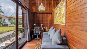 A room with wooden walls, a grey sofa, a desk with a chair, a hanging light fixture, and a large window with a view of houses and greenery. - Lumbung Mas Villa by Betterplace (Ubud)