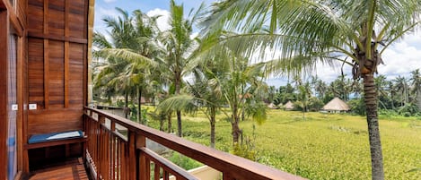 A wooden balcony with a bench, overlooking a lush green field with palm trees and a thatched-roof structure in the distance.