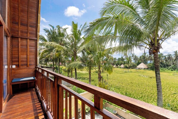 A wooden balcony with a bench, overlooking a lush green field with palm trees and a thatched-roof structure in the distance.