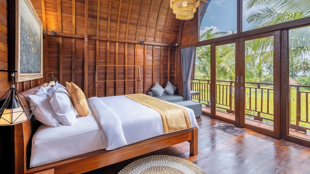 A wooden room with a bed, a hanging light fixture, and a view of palm trees. - Lumbung Mas Villa by Betterplace (Ubud)