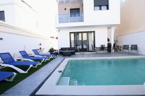 Un área de piscina moderna al aire libre con sillas de descanso, una zona de estar y una escalera que lleva a un balcón.