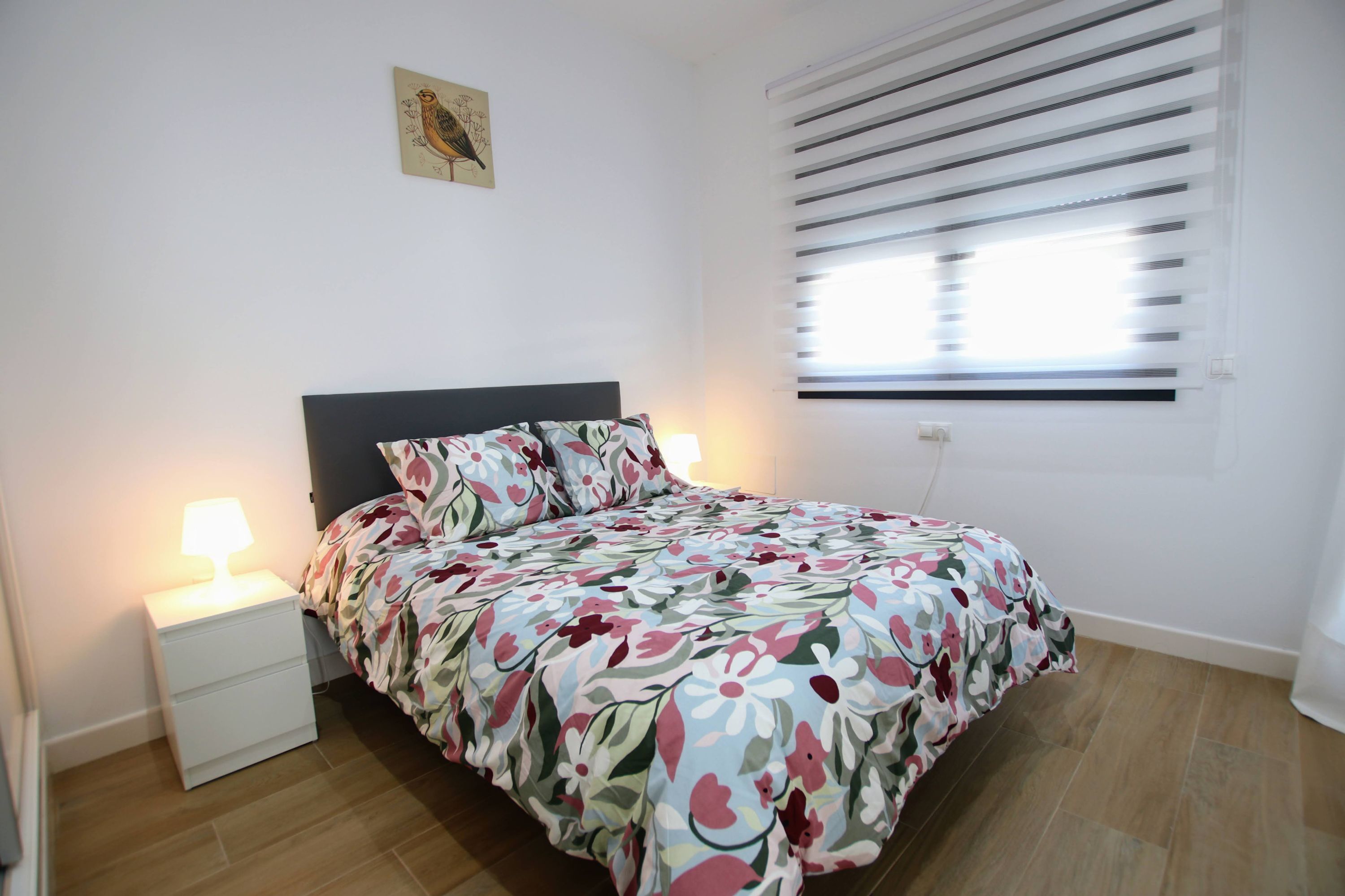 A bedroom with a bed, nightstands, a wall-mounted lamp, a window with blinds, and a wall decoration.