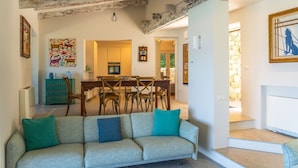 A living room with a sofa, a dining area with a table and chairs, and a kitchen in the background. - Villa Onda Celeste - Sardinia Unlimited (Costa Paradiso)