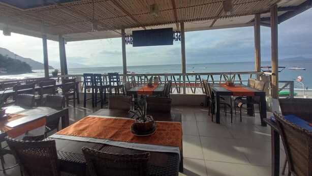 Un restaurante al aire libre con mesas y sillas, una pantalla de televisión y vista al océano.
