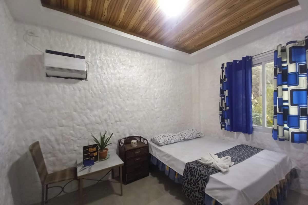 Una habitación pequeña y limpia con dos camas individuales, un escritorio, una silla y una ventana con cortinas azules.