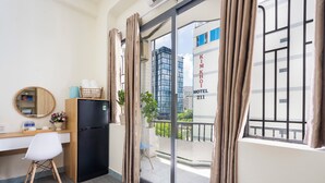 A hotel room with a desk, a black refrigerator, a small chair, a round mirror, and a view of the city through large windows. - Phan Hotel (Ho Chi Minh City)