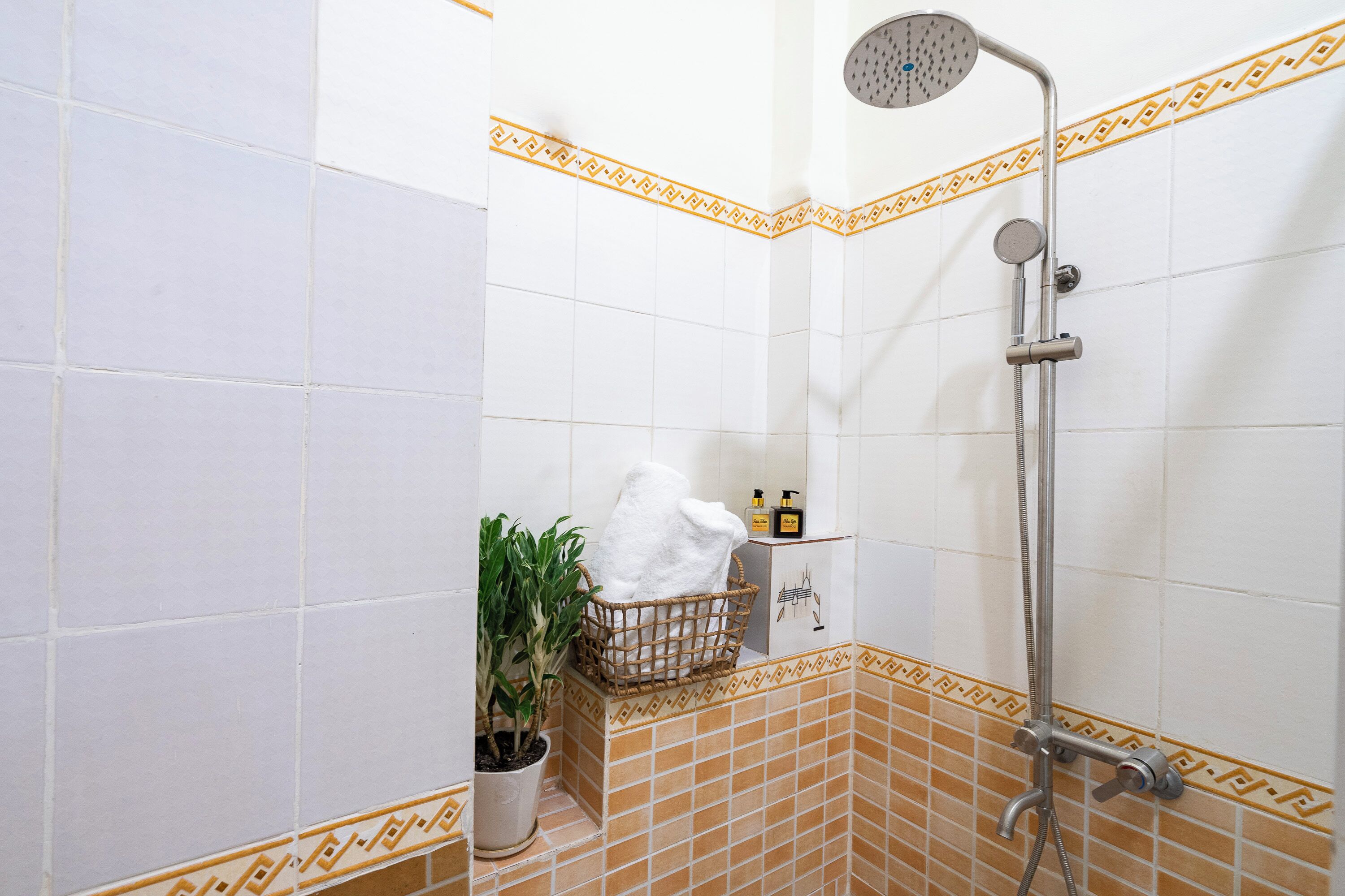 Ein Badezimmer mit weißen und orangefarbenen Fliesen, einer Dusche, einem Handtuchhalter, einer Topfpflanze und Pflegeprodukten.